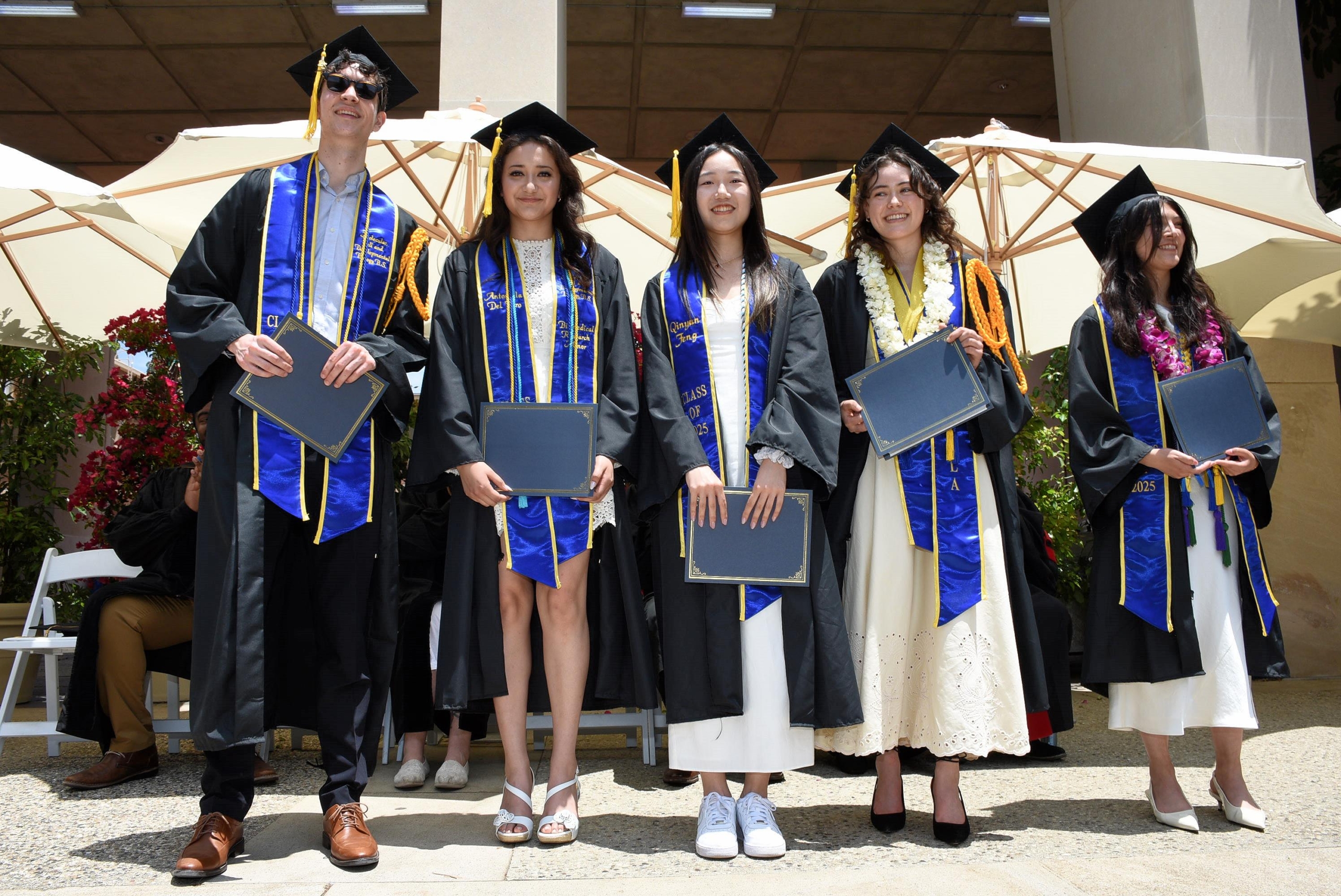 Four UCLA graduates in blue and gold sashes standing together on campus.